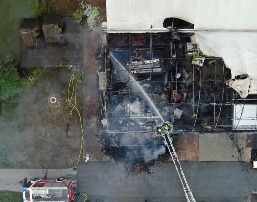 Eine Luftaufnahme eines brennenden Gebäudes, bei dem Feuerwehrleute eine Leiter benutzen, um die Flammen zu löschen. Rauch und Wassernebel sind sichtbar.