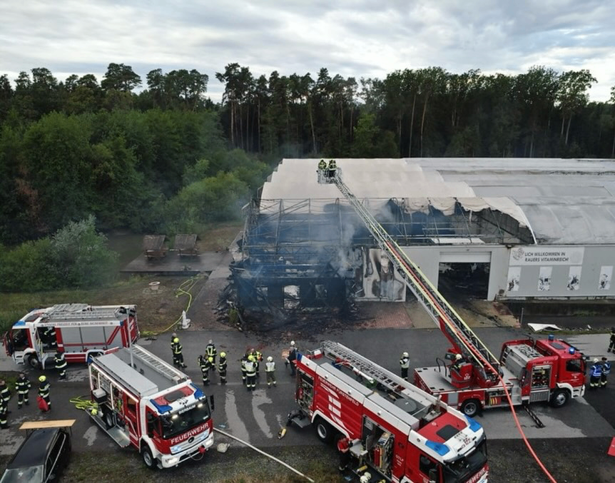 Ein Löscheinsatz ist im Gange bei einem ausgebrannten Gebäude mit mehreren Feuerwehrfahrzeugen und Feuerwehrleuten. Das Gebäude hat ein großes weißes Dach und ist von Bäumen umgeben.