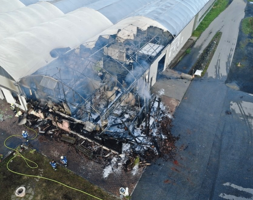 Eine Luftaufnahme eines schwer beschädigten Industriegebäudes mit aufsteigendem Rauch aus den Trümmern, Feuerwehrleute sind am Boden anwesend.