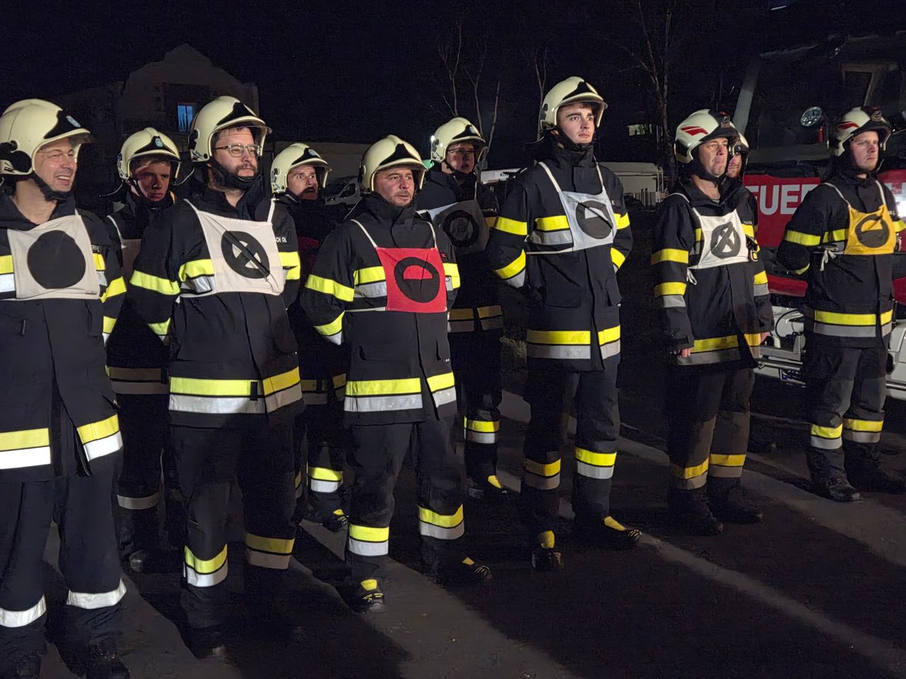 A group of firefighters in full uniform stands on a street at night. They wear helmets, glasses, and reflective vests. Buildings are visible in the background.
