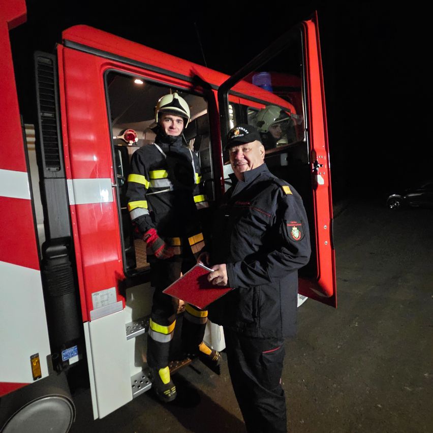 Two firefighters in full gear, one holding a clipboard, stand in front of a red fire truck with its door open at night.