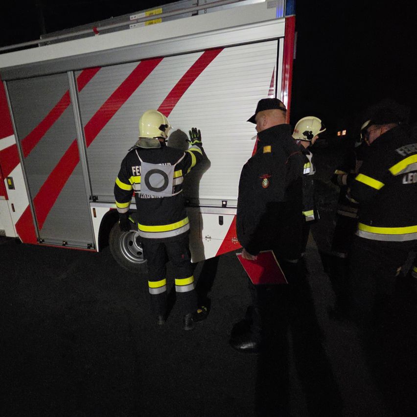 Firefighters are gathered around a fire truck, some are wearing helmets and gloves. One firefighter is touching the side of the truck.