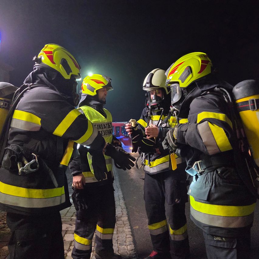 Four firefighters in full gear stand in a line, wearing yellow helmets, holding equipment, and having a conversation. One of them is holding a tool. They are outside at night.