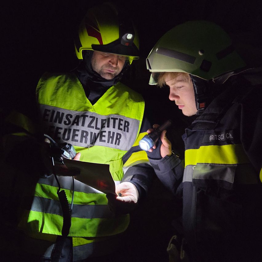 Two firefighters are looking at a piece of paper in a dark room, one holding a flashlight. The one on the left wears a yellow vest reading 'EINSATZLEITER FEUERWEHR'.