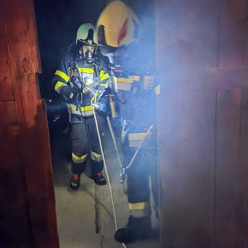 Two firefighters in full gear enter a smoky room. One holds a rope, and both wear helmets. The room has wooden walls and a cracked floor.