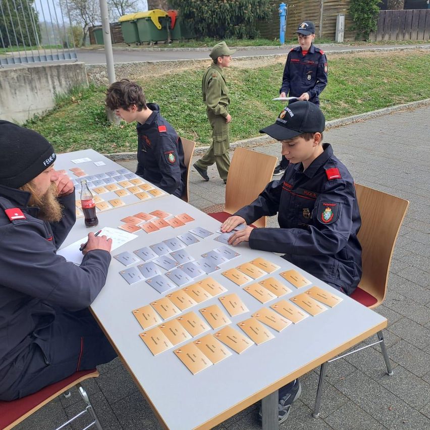 Several people in uniforms sit around a table filled with cards. One person is writing on paper while another examines a card. A uniformed person walks in the background.