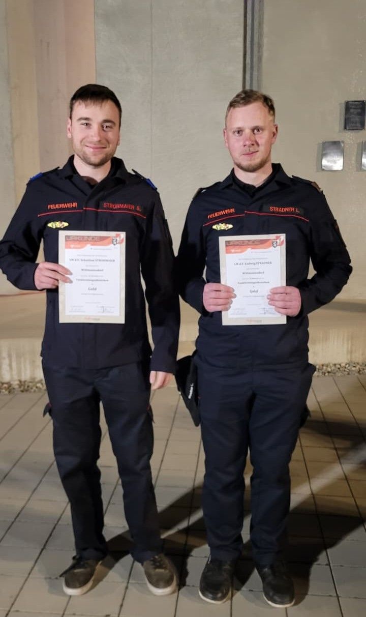 Zwei Feuerwehrleute stehen zusammen und halten Urkunden in den Händen, lächeln für ein Foto. Sie tragen Uniformen mit Aufnähern, die mit 'Feuerwehr' und Namen beschriftet sind. Die Urkunden sind golden.