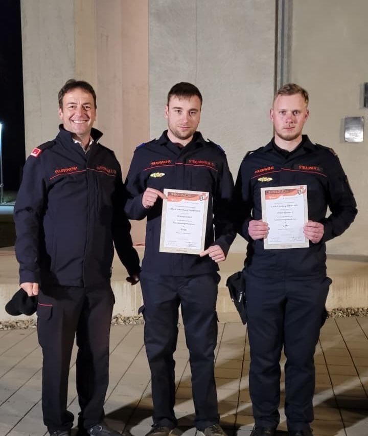 Drei Männer in Marineuniformen posieren mit Urkunden in der Hand für ein Foto. Der Mann links lächelt, während die anderen ernst aussehen. Sie stehen auf einem gefliesten Boden vor einer Betonwand.