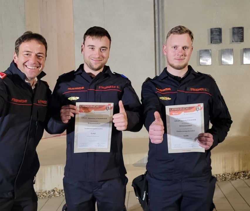 Drei Feuerwehrleute in Uniform posieren mit einem Zertifikat für ein Foto, lächeln und geben Daumen hoch. Hinter ihnen hat eine Wand Plaketten.