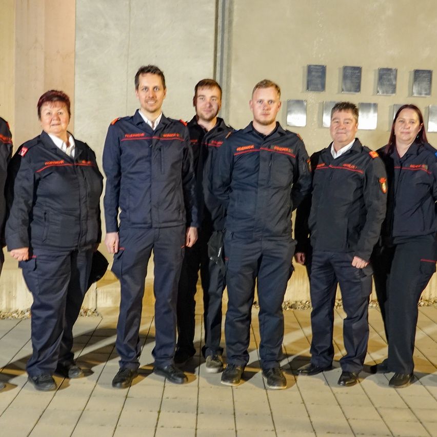 Eine Gruppe von Feuerwehrleuten in Uniform steht in einer Reihe und lächelt für ein Foto vor einer Wand mit Plaketten.