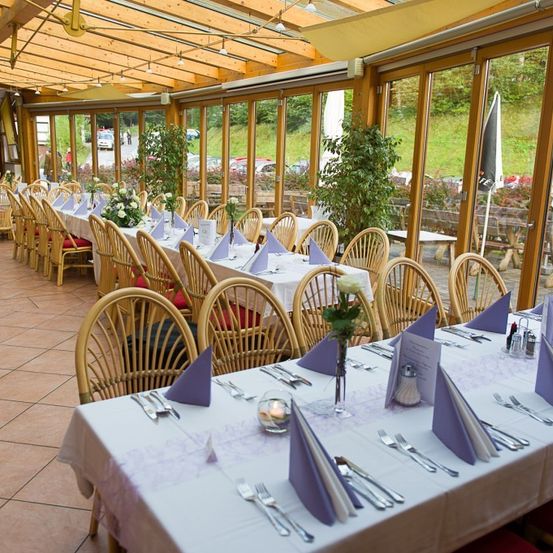 Ein gut beleuchtetes Innenrestaurant mit Holztischen und Stühlen, geschmückt mit weißen Tischdecken und lila Servietten. Der Raum hat große Glasfenster, die einen Blick auf die Außenlandschaft bieten.