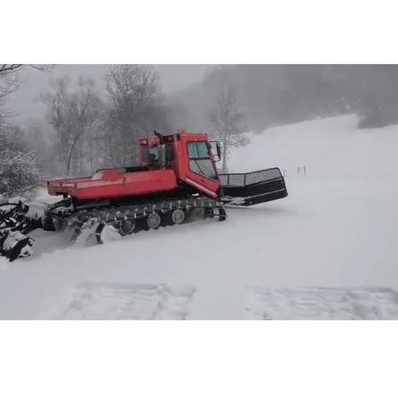 Ein roter Schneepflug mit Raupenketten räumt einen verschneiten Weg. Er befindet sich in einem Gebirgsgebiet mit nebliger Atmosphäre.