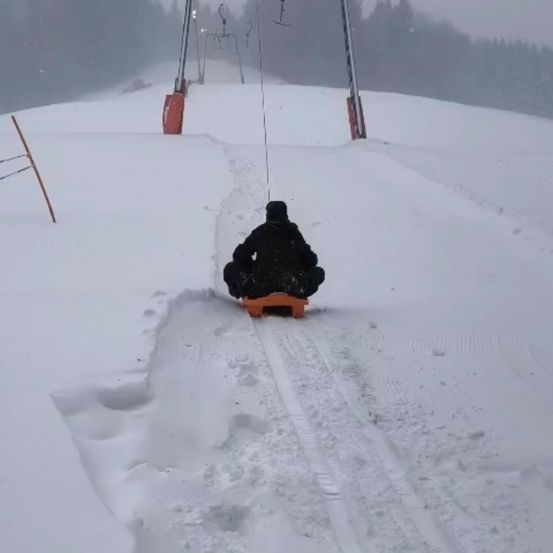 Eine Person fährt auf einem Schlitten einen verschneiten Hang hinunter, umgeben von Schnee und Skiliftstangen.