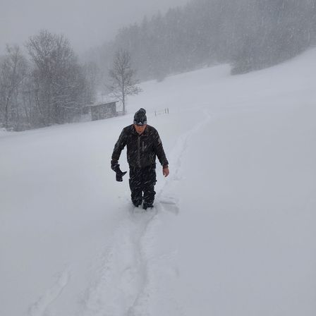Ein Mann geht in einem verschneiten Gebiet. Er ist in einem schwarzen Jacken-, Hosen- und Hutoutfit gekleidet. Er schaut nach unten und scheint sich auf etwas zu konzentrieren. Bäume und ein kleines Haus sind im Hintergrund.