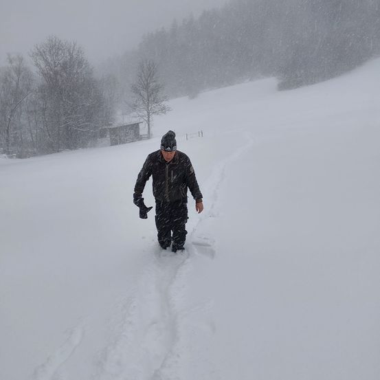 Ein Mann geht in einem verschneiten Gebiet. Er ist in einem schwarzen Jacken-, Hosen- und Hutoutfit gekleidet. Er schaut nach unten und scheint sich auf etwas zu konzentrieren. Bäume und ein kleines Haus sind im Hintergrund.