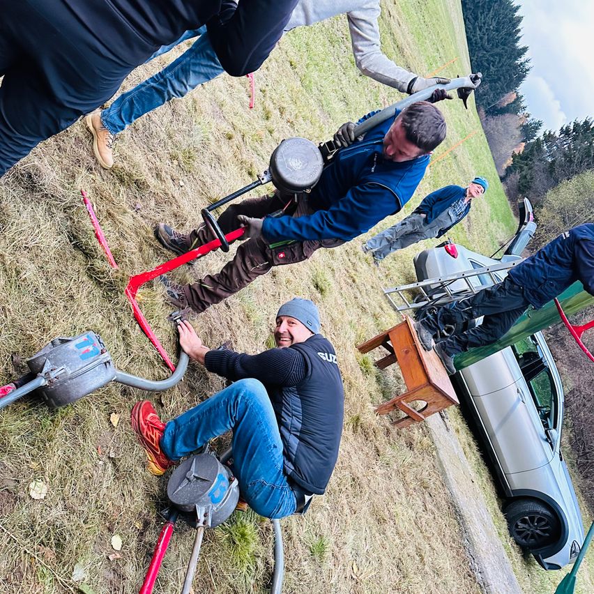 Eine Gruppe von Menschen arbeitet auf einem Grasfeld. Ein Mann kniet und arbeitet an einer Maschine, während andere mit Werkzeugen stehen. Ein silbernes Auto ist in der Nähe geparkt.