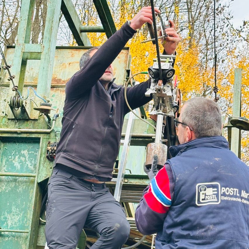 Zwei Männer sind auf einem Fahrzeug, wahrscheinlich einem Versorgungsfahrzeug. Ein Mann klettert eine Leiter hinauf und hält sich an einer Maschine fest. Der andere Mann, der eine Brille trägt, steht neben ihm. Beide sind in Arbeitskleidung gekleidet. Das Fahrzeug hat rostiges Metall und ist von Bäumen umgeben.