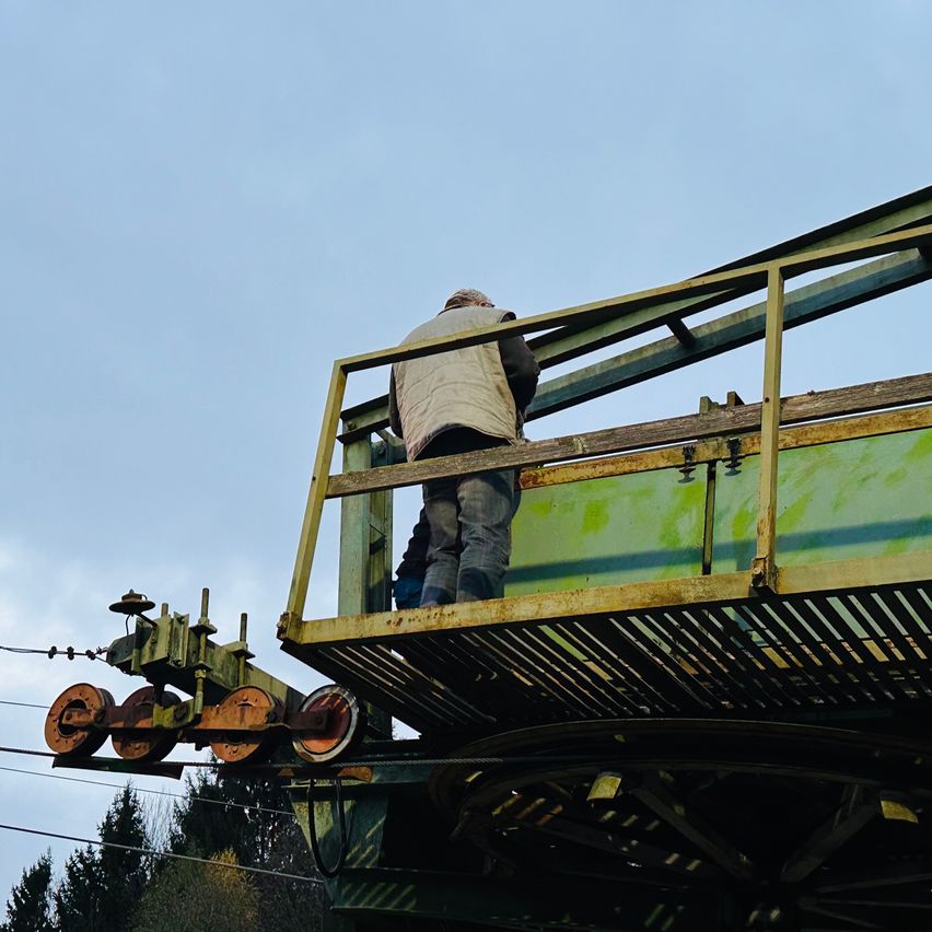 Ein Mann steht auf der Kante einer rostigen grünen Metallplattform einer Seilbahn und blickt nach unten.