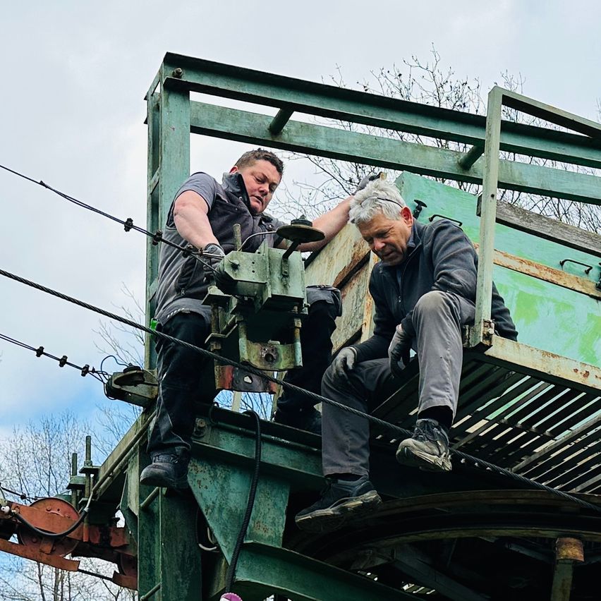 Two men are working on a green metal structure. One is sitting, and the other is standing, both wearing gloves. The sitting man is smiling.