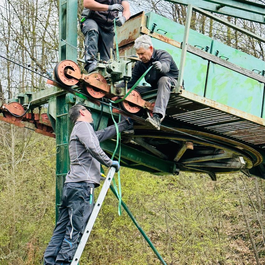 Three men are working on a green structure with wheels and cables. One man is on a ladder, another is sitting, and a third is standing on the structure.