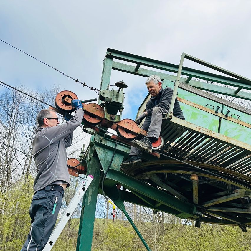 Two men in work gear are working on a cable car. One is climbing up a ladder, and the other is seated on a platform, both under a cloudy sky.