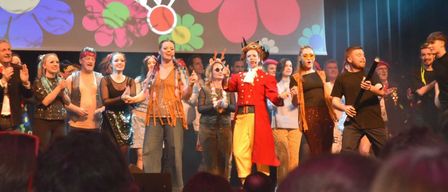 Eine Gruppe von Darstellern auf der Bühne mit einem bunten Hintergrund, auf dem 'Faschings Show' steht. Sie sind in verschiedenen Kostümen gekleidet. Einer trägt einen roten Mantel und gelbe Hosen. Im Vordergrund sind Zuschauer.