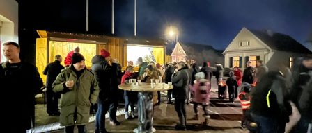 Eine Gruppe von Menschen versammelt sich bei Nacht draußen um einen Tisch, vielleicht bei einem Festival. Ein Mann in einem grünen Mantel hält einen Becher. Straßenlaternen beleuchten das Gebiet.