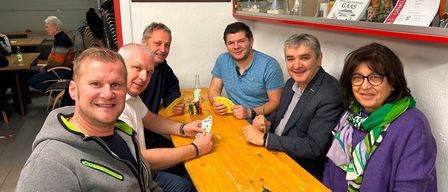 Eine Gruppe von Männern sitzt an einem Holztisch mit Würfeln und Karten und lächelt. Ein Trophäenschrank mit Bechern und einem Schild dahinter. Ein Gemälde eines Hauses an der Wand.