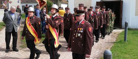 Eine Gruppe von Männern in Militäruniformen und Helmen geht auf einem Ziegelweg, eine Fahne haltend, vor einem Gebäude mit Zuschauern an der Seite.