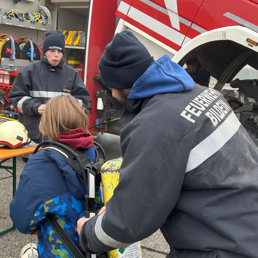Ein Mann hilft einem Kind, ein Atemschutzgerät eines Feuerwehrmanns anzupassen, während ein weiterer Feuerwehrmann in der Nähe eines Feuerwehrwagens steht.