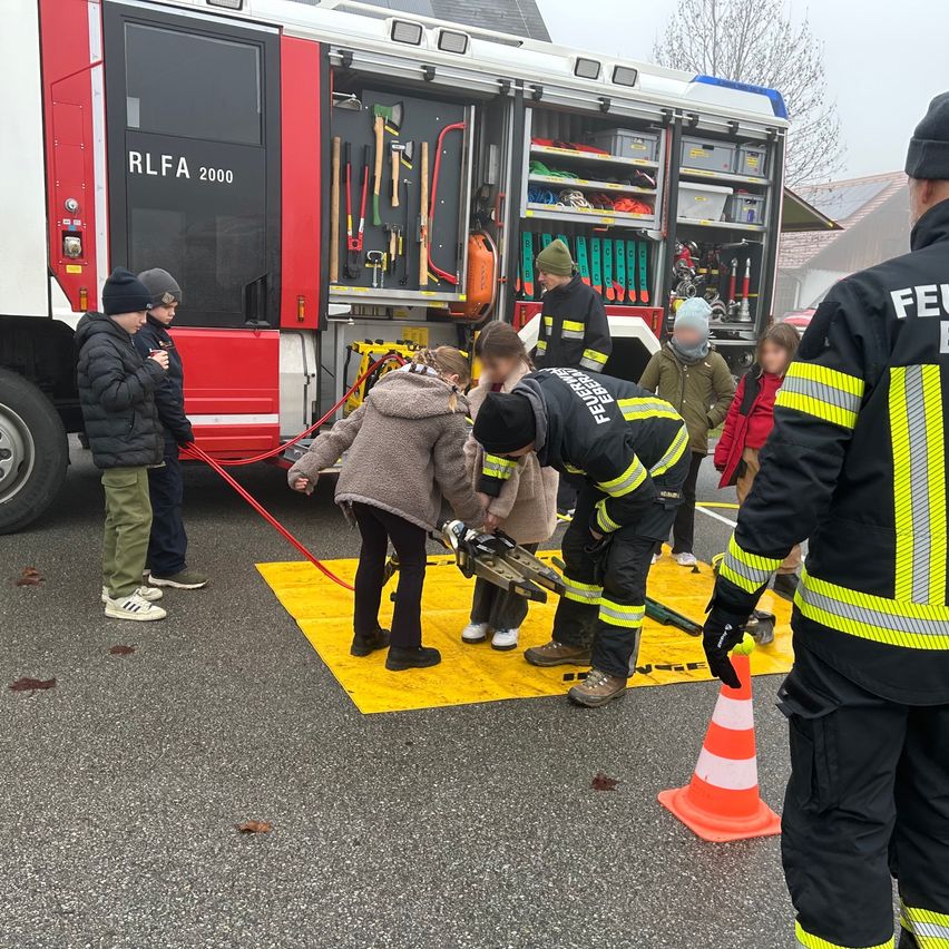 Feuerwehrleute demonstrieren Ausrüstung für Kinder. Der Feuerwehrwagen ist offen, Werkzeuge sind sichtbar. Eine gelbe Matte liegt zum Schutz auf dem Boden.
