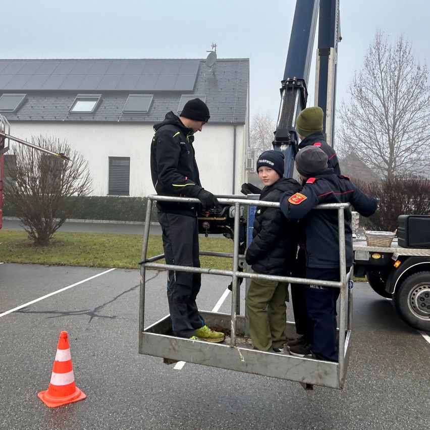 Ein Mann steht in einem Metallkorb mit drei Kindern auf einem Parkplatz. In der Nähe steht ein Kran und ein Gebäude mit Solarpanelen.