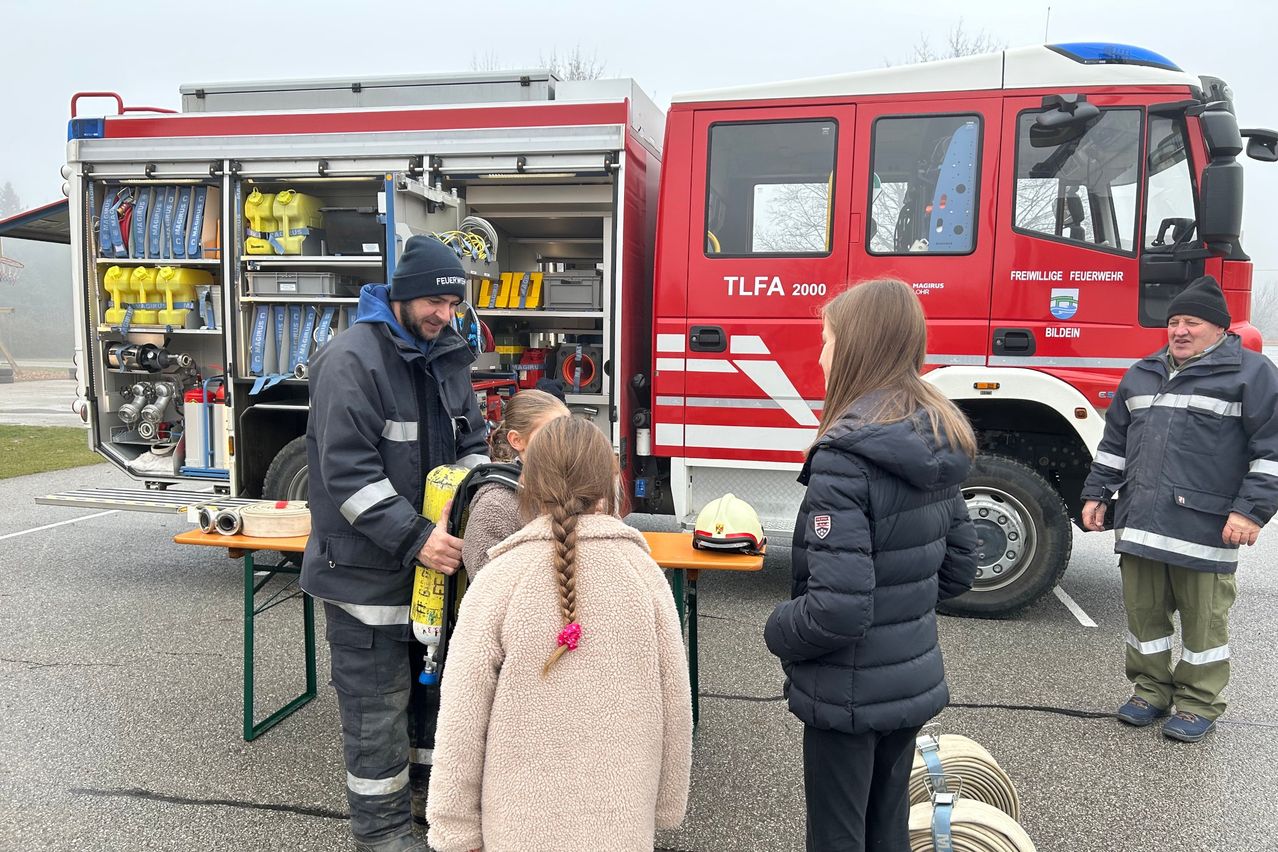 Ein Feuerwehrmann demonstriert zwei Kindern vor einem Feuerwehrauto eine Sauerstoffflasche.