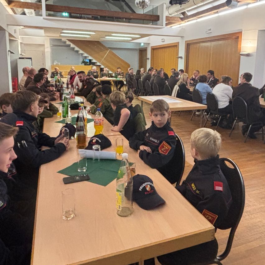 Eine große Gruppe von Menschen in Uniformen sitzt an einem langen Tisch mit verschiedenen Gegenständen. Einige sitzen auf Stühlen, andere stehen. Auf dem Tisch befinden sich Gläser, Flaschen und ein Telefon.