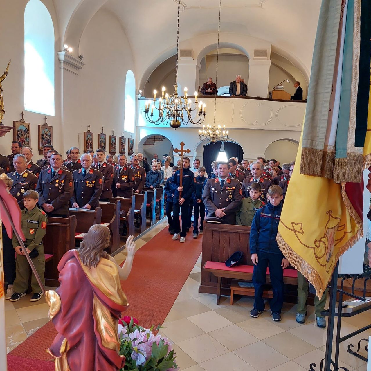 A group of military personnel and children stand in a church with a statue, flags, and chandeliers. Some stand by the pews while others are on the aisle.
