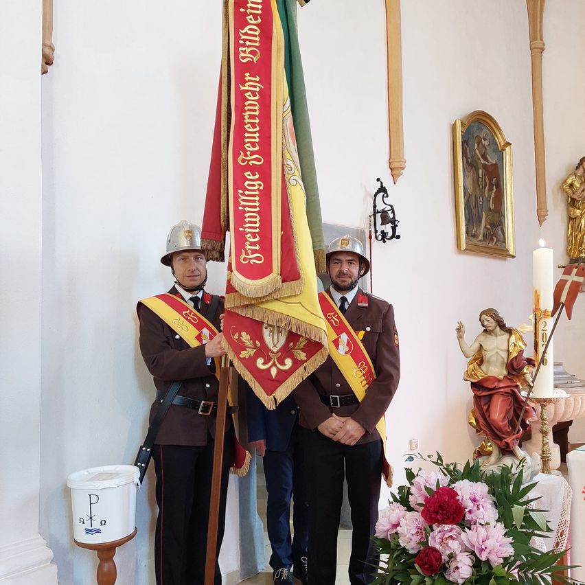 Two men in military uniforms stand in front of a white wall, holding a flag and a flagpole. A bucket with the letters PKA is on the left. A white candle is on the right, and a statue of Jesus is behind it.