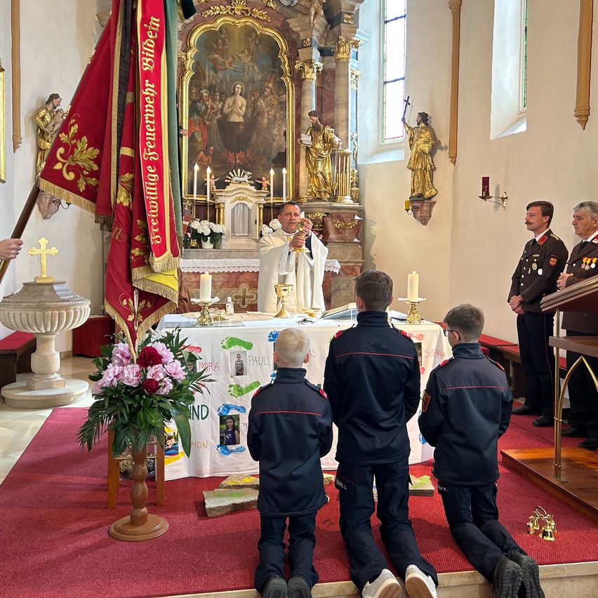 A priest leads a religious ceremony in a church, with four boys in uniforms kneeling before the altar. A flag and flowers adorn the area, and two men in military uniforms stand nearby.