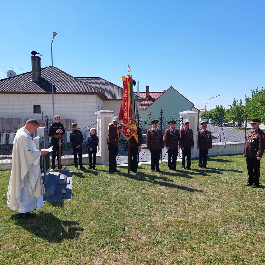 A priest in a white robe leads a solemn ceremony in front of a memorial with military officers standing in formation.