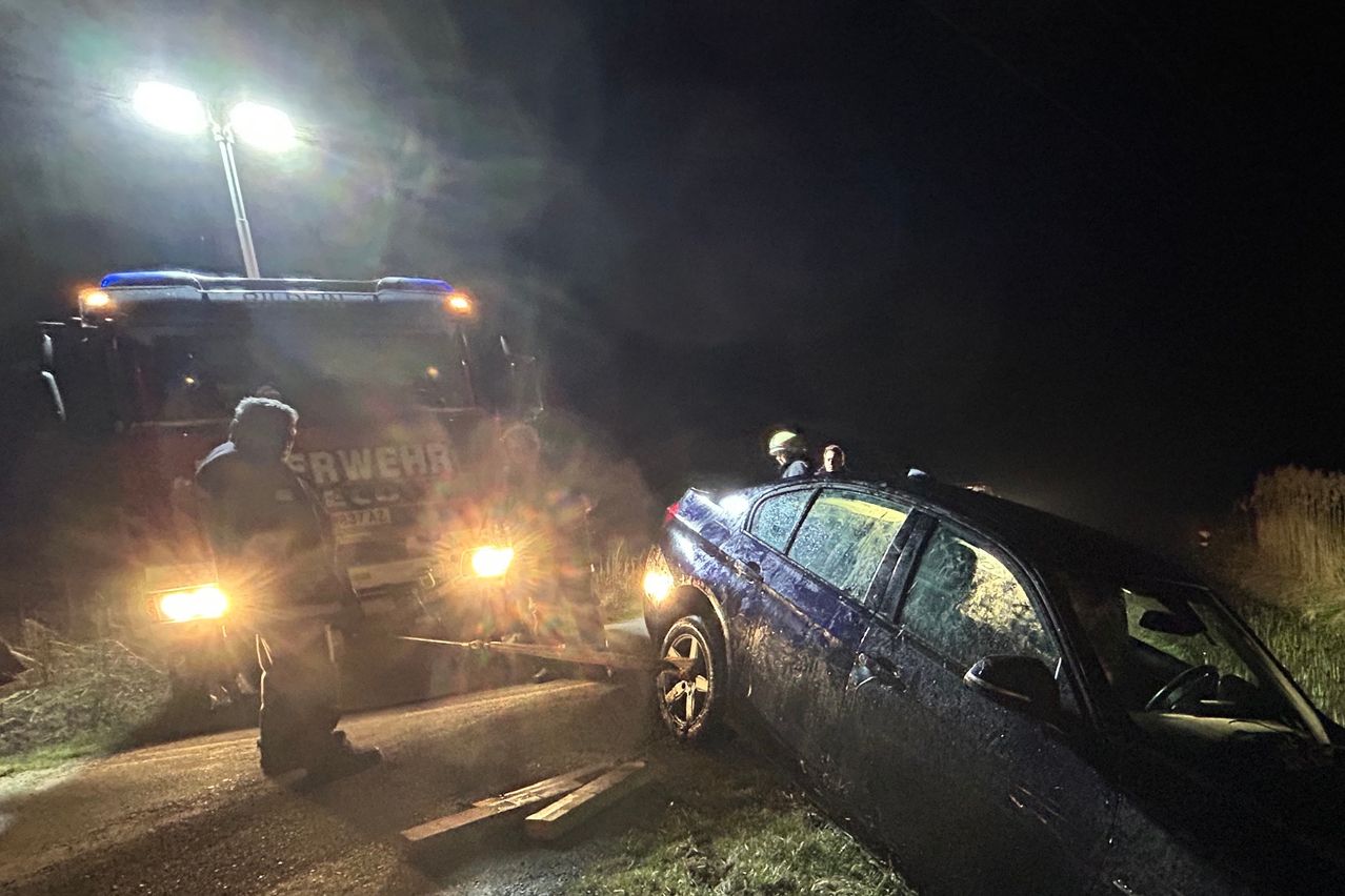 A fire truck with illuminated headlights is rescuing a dark blue car that is stuck on the side of the road.