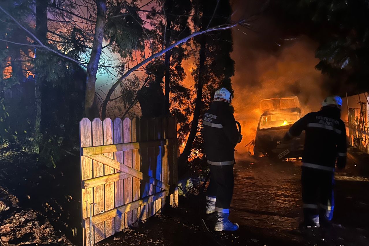 A firefighter stands near a burning vehicle, the fire behind him illuminating the night sky. Trees and a wooden fence are in the foreground.