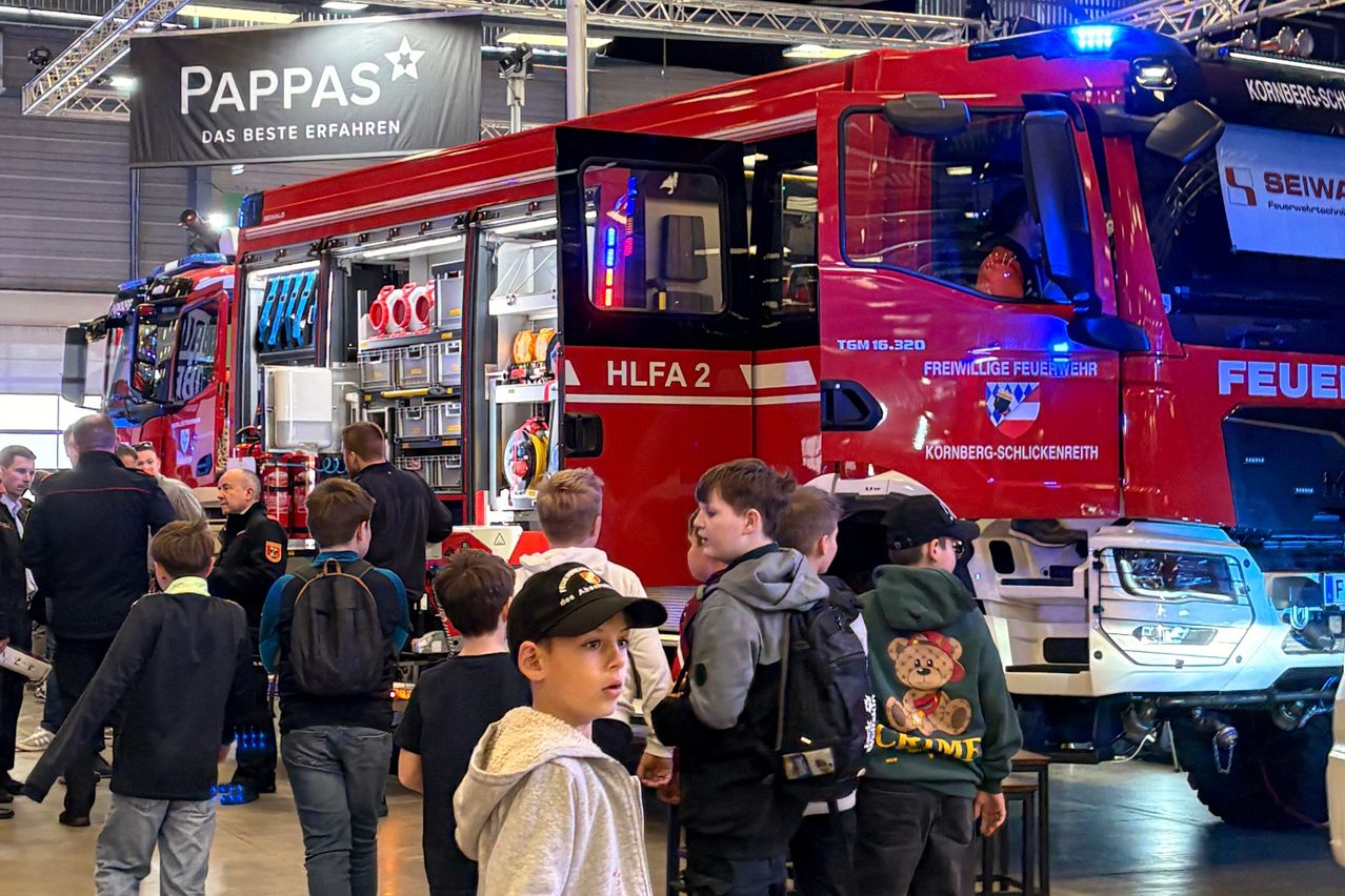 Eine Gruppe von Kindern steht vor einem roten Feuerwehrauto mit der Aufschrift HLFA 2. Sie tragen alle Jacken und haben Rucksäcke dabei. Einer von ihnen trägt einen Hut. An der Wand befindet sich ein Schild mit der Aufschrift APPAS.