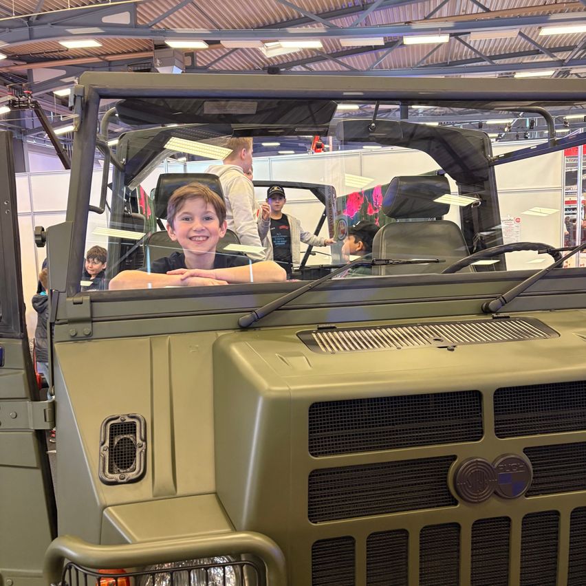 Ein junger Junge sitzt lächelnd auf dem Fahrersitz eines Militärfahrzeugs. Im Hintergrund sind mehrere Personen zu sehen, und das Fahrzeug befindet sich in einem großen Innenbereich.