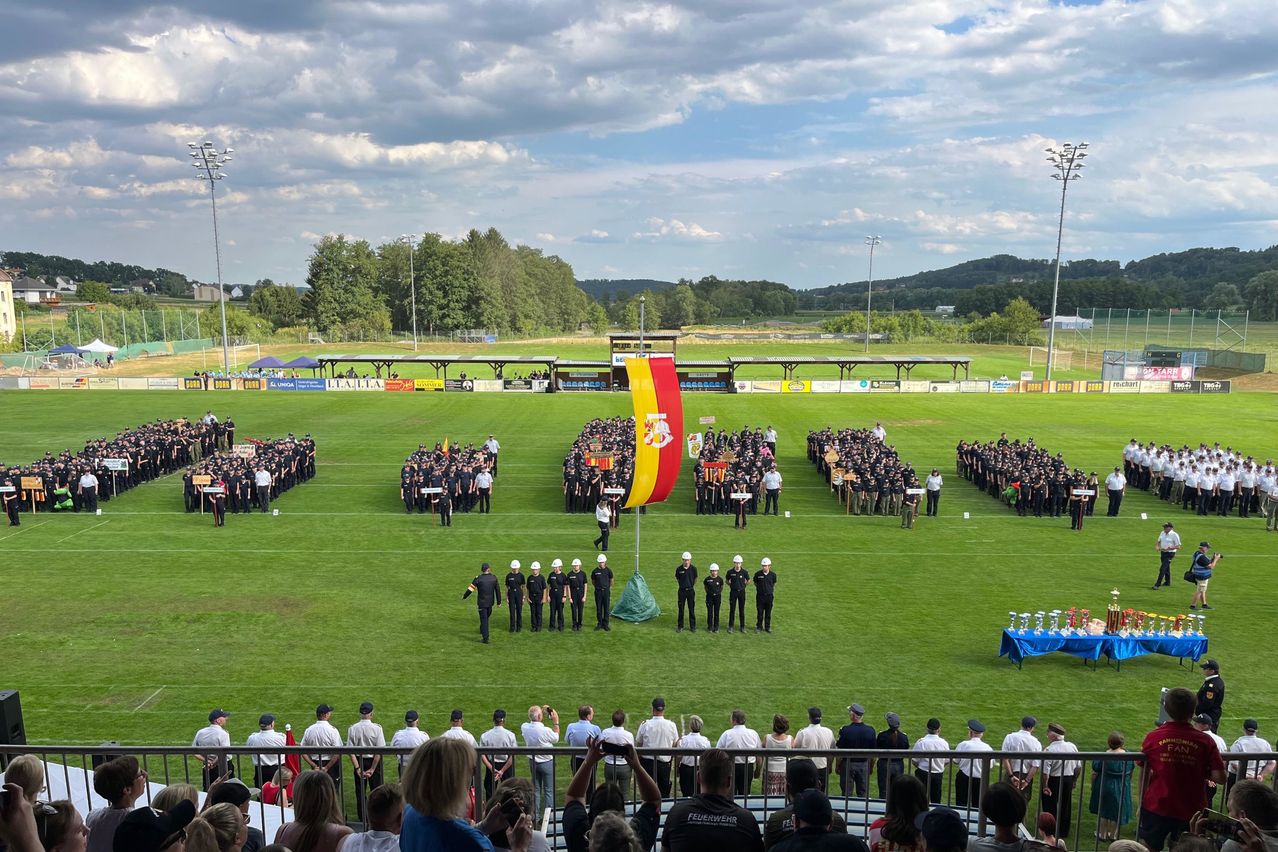 Eine große Gruppe von Menschen in Uniform steht auf einem Rasenfeld, mit einer Flagge in der Mitte. Zuschauer sitzen auf den Tribünen, und es gibt Banner und Flaggen im Hintergrund.