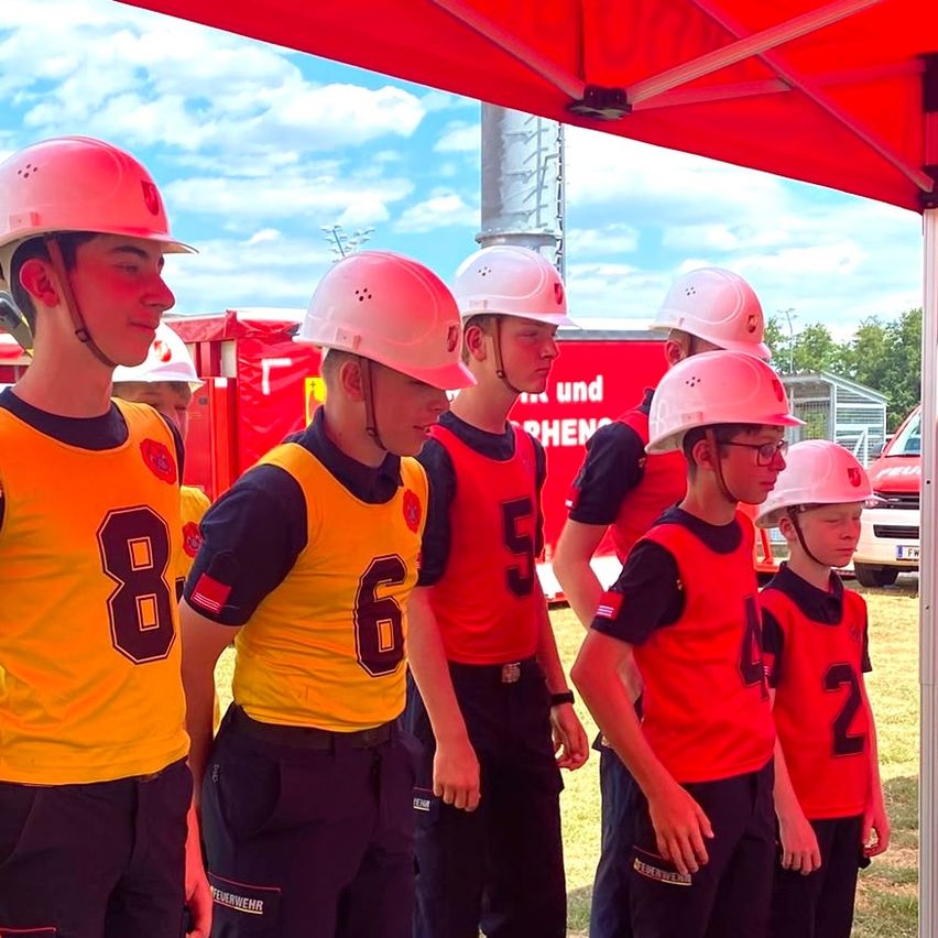 Eine Gruppe junger Feuerwehrleute in Uniform, jeder mit einem nummerierten Helm, steht unter einem roten Zelt. Im Hintergrund ist ein roter Feuerwehrwagen und ein rotes Gebäude zu sehen.