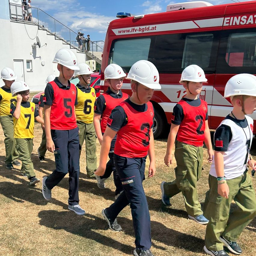Eine Gruppe von Kindern in roten und gelben Uniformen mit Nummern darauf geht vor einem roten Feuerwehrwagen.