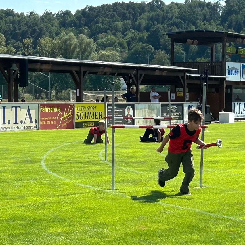 Ein junger Junge läuft und springt über eine Hürde auf einem Rasenfeld mit einem Pavillon im Hintergrund.