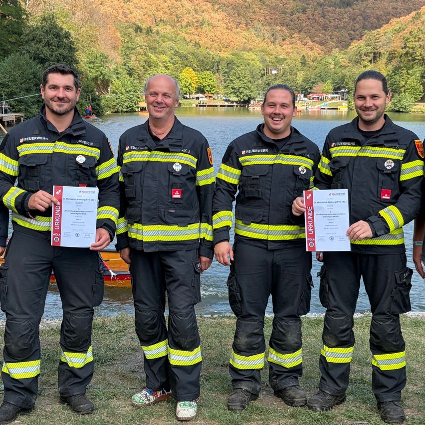 Vier Feuerwehrleute in Uniform stehen am See, lächeln und halten Urkunden. Hinter ihnen spiegelt der See Bäume und ein entferntes Boot wider.