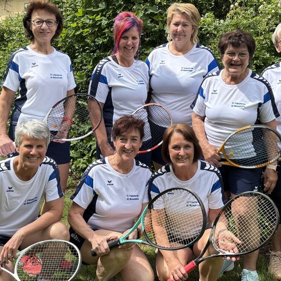 Eine Gruppe von Frauen in einheitlichen Trikots posiert mit Tennisschlägern in einem Rasenbereich, mit Pflanzen und einem Gebäude im Hintergrund.