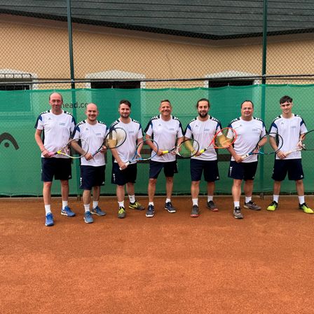 Sechs Männer in weißen Shirts und schwarzen Shorts stehen auf einem Tennisplatz mit Schlägern, lächeln und posieren für ein Foto.