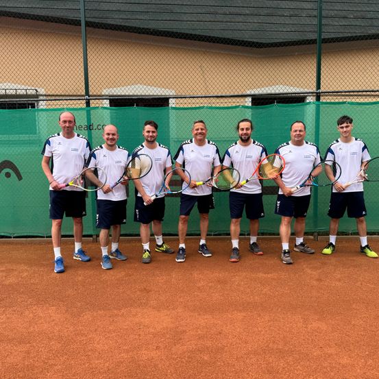 Sechs Männer in weißen Shirts und schwarzen Shorts stehen auf einem Tennisplatz mit Schlägern, lächeln und posieren für ein Foto.